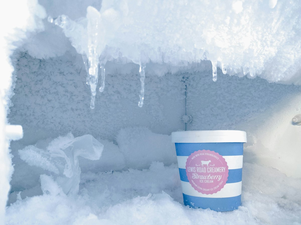 Heavy frost and icicles covering the interior walls of a home freezer beside a tub of ice cream