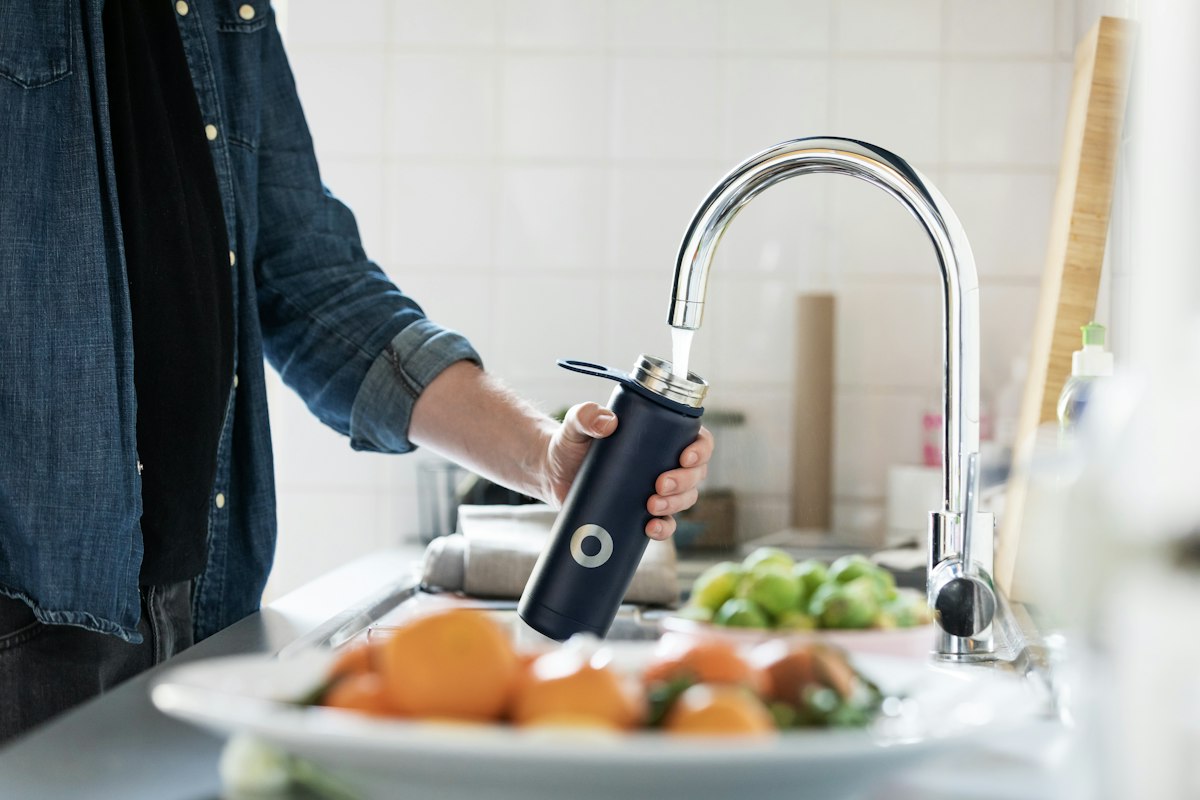 A person filling a dark bottle with running water from a chrome kitchen faucet
