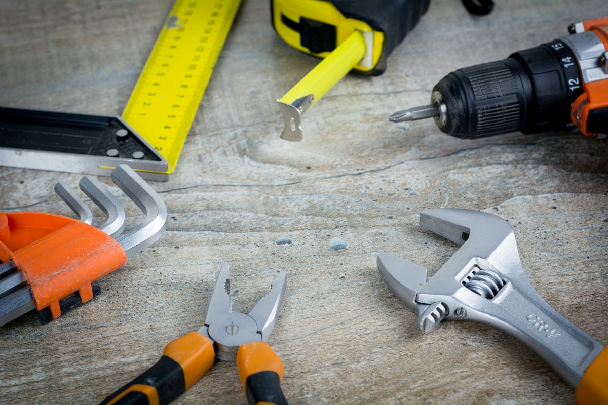 A cordless drill, adjustable wrench, pliers, hex keys and a folding rule laid out on a workbench