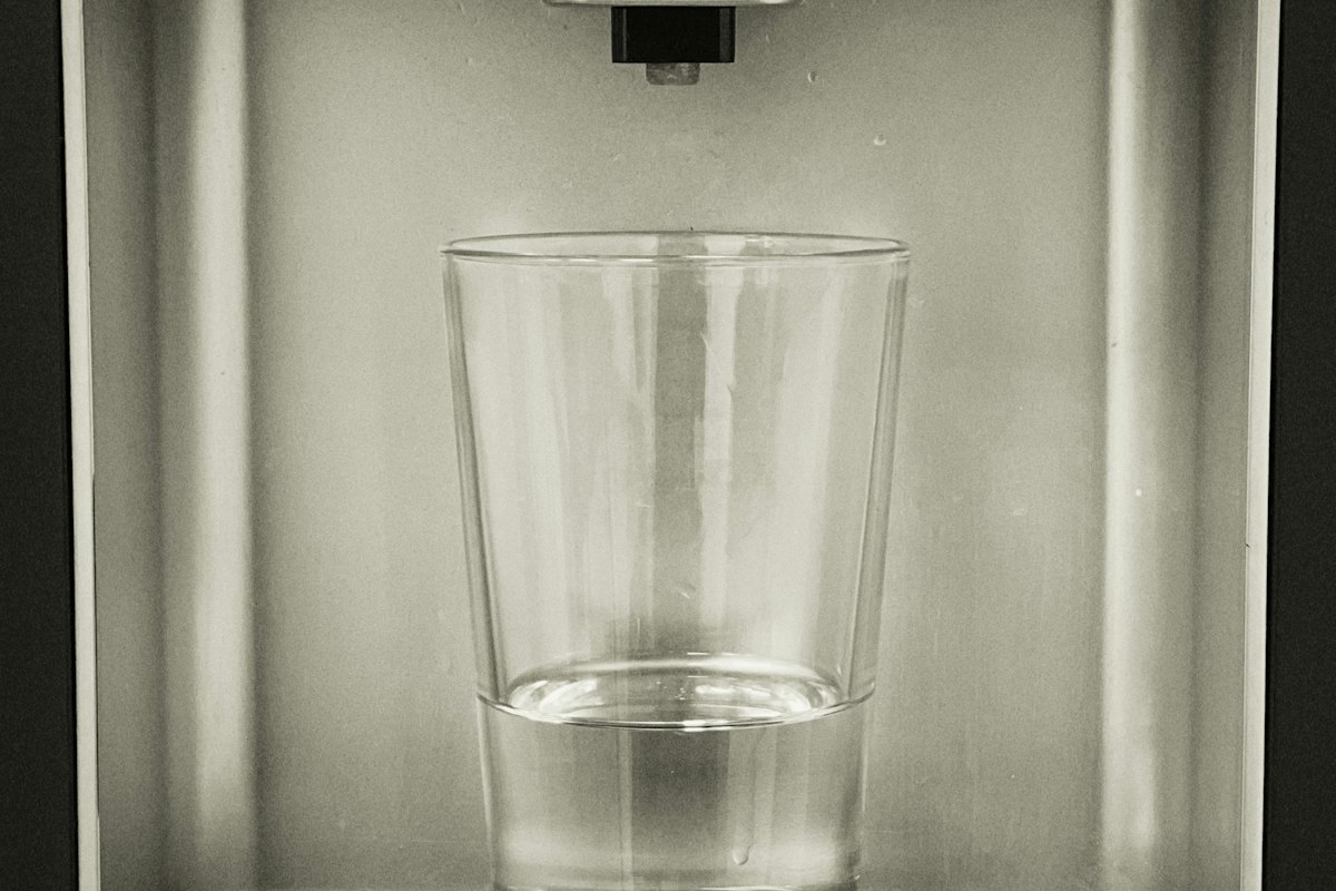 A clear glass half full of water sitting in a stainless steel refrigerator water dispenser